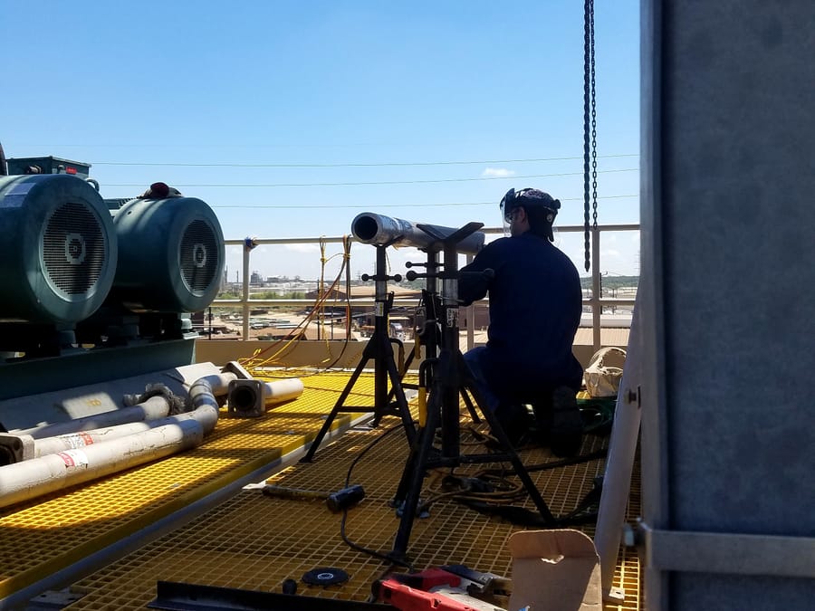 A technician working on a pipe spool on jack stands on an elevated grating platform, the plant behind.