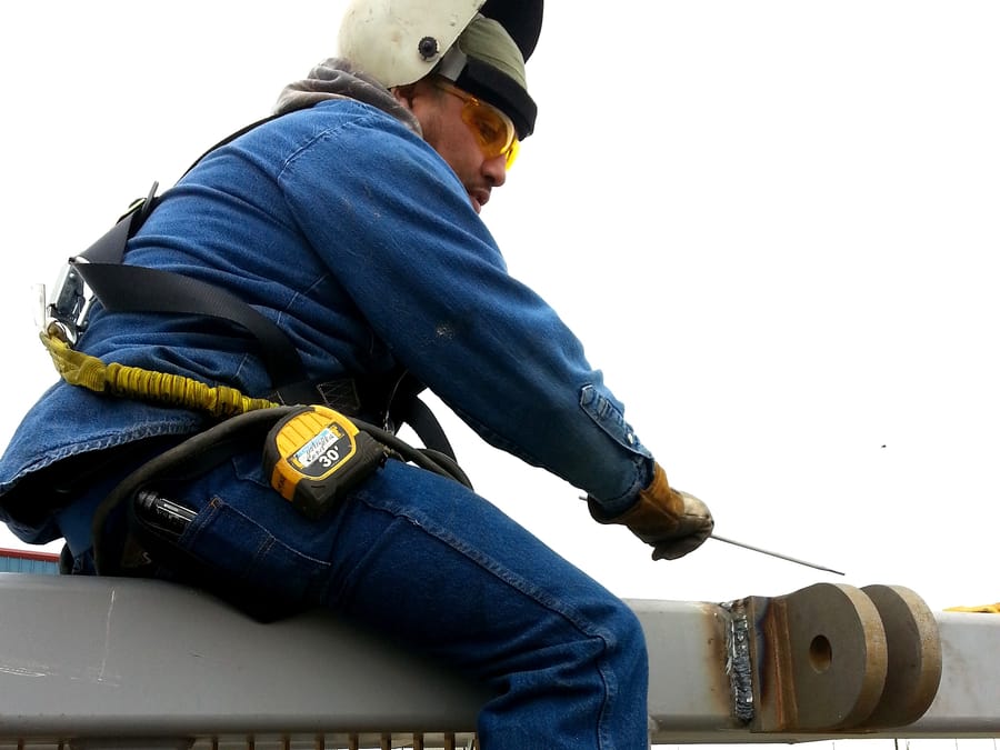 A welder in a fall-arrest harness working at height on a structural member, the finished weld alongside.
