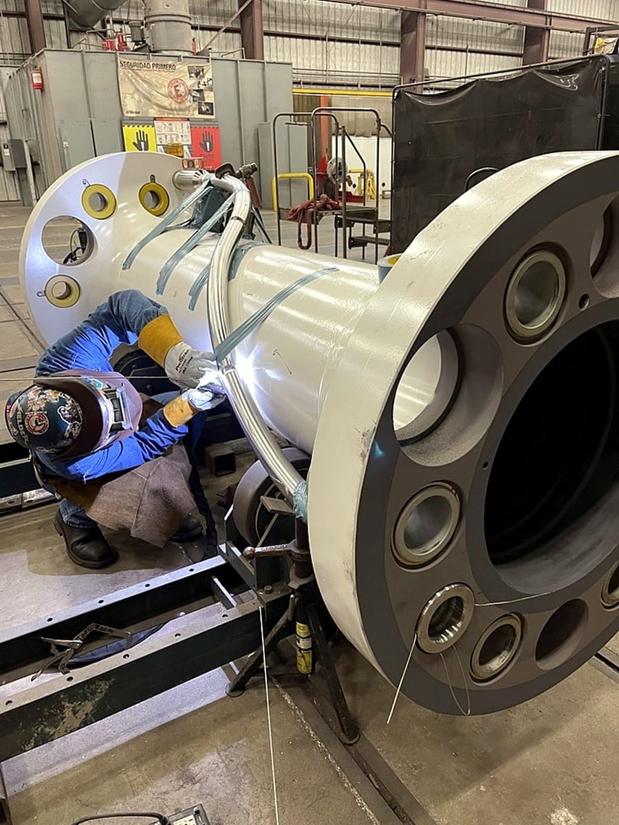 A welder working on a large flanged cylindrical assembly supported on stands.