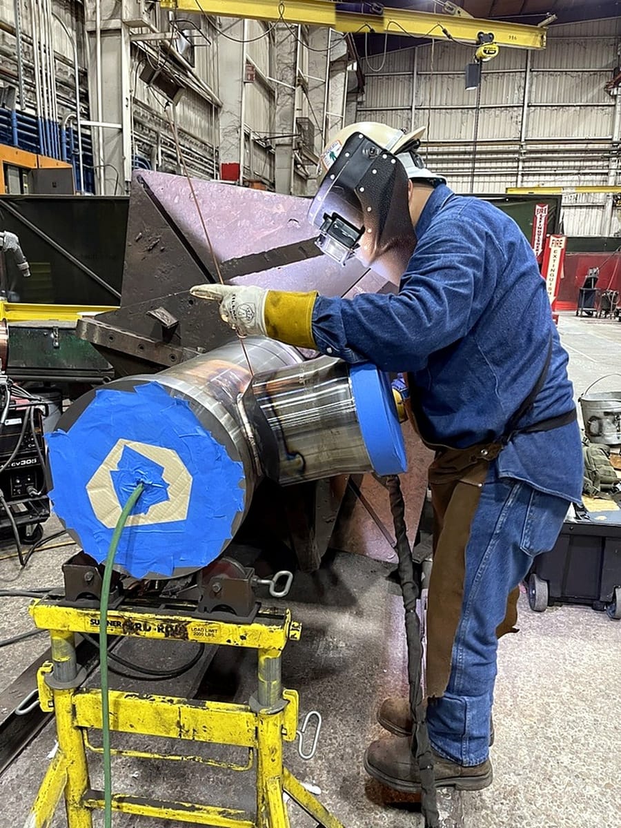 A welder running a pass on a shaft held in a welding positioner.