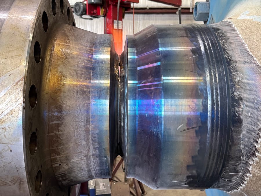 A heavy flanged body with machined and heat-tinted surfaces, a prepared weld groove at the centre and a weld build-up band at the right.