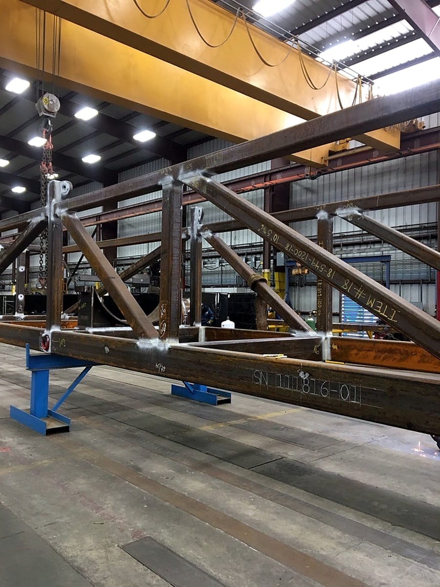 A long structural frame under fabrication, supported on stands beneath the crane.
