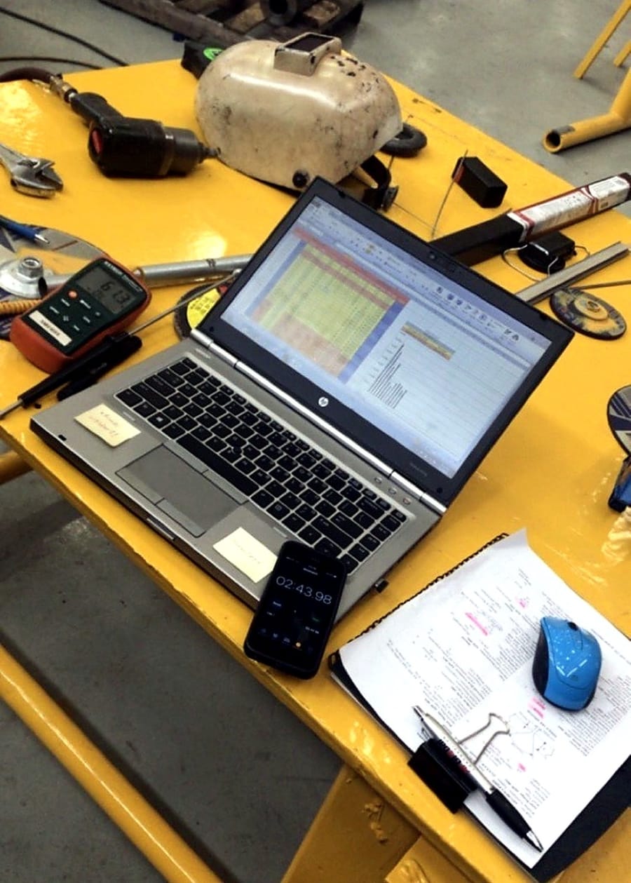 A laptop logging welding parameters at the work station, procedure sheets alongside.